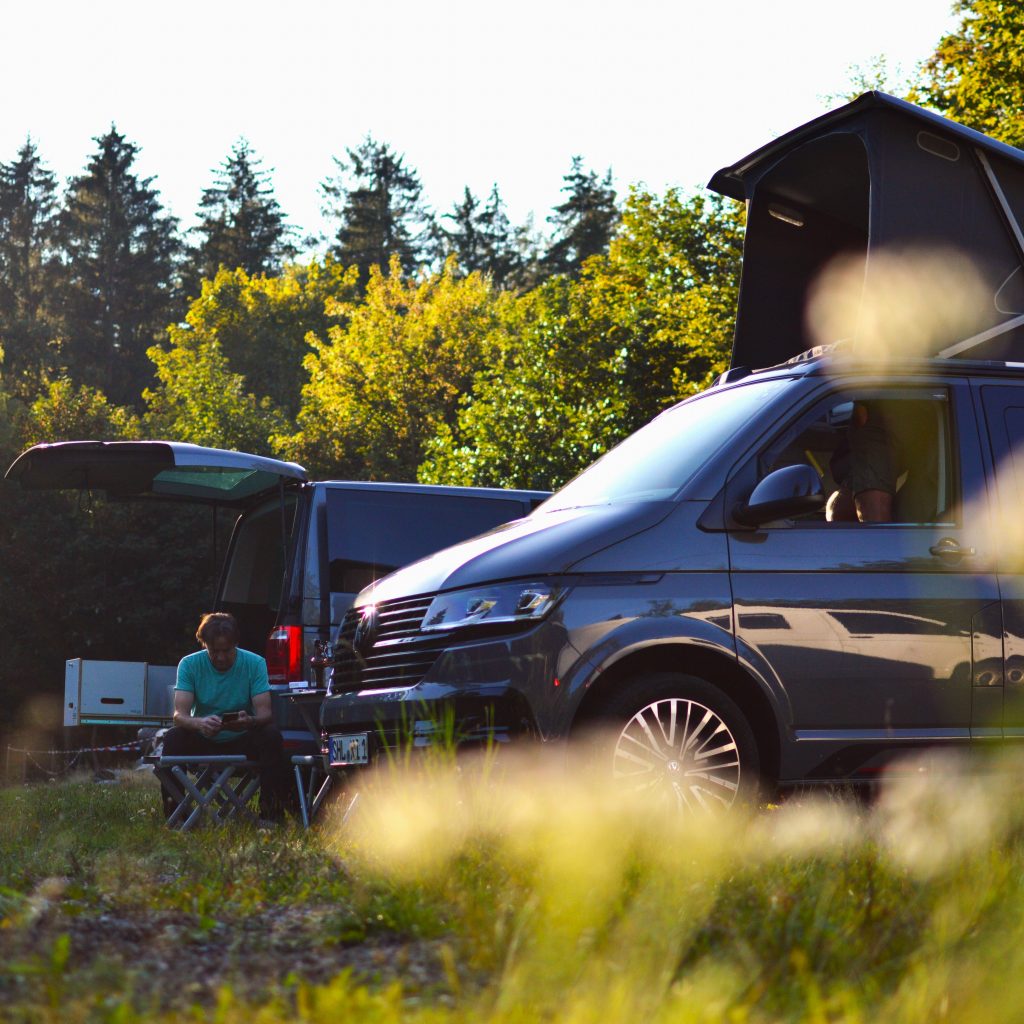 Zwei Camper stehen auf einer Wiese vor einem Waldrand.
