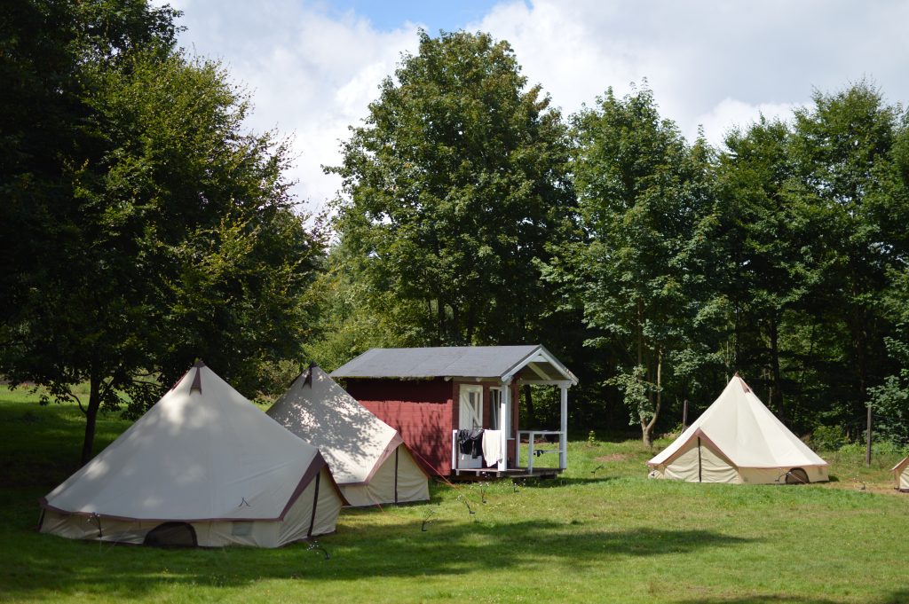 Zeltlager mit mehreren weißen Tipi-Zelten auf einer grünen Wiese vor einem roten Holzhaus am Waldrand.